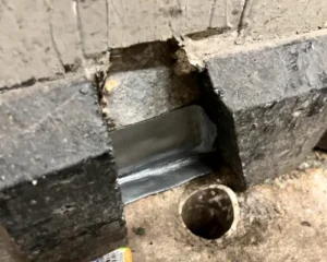 High-angle shot above an open drainage pipe, showcasing damaged concrete baseboard reinforced with a silver flex sealant patch.